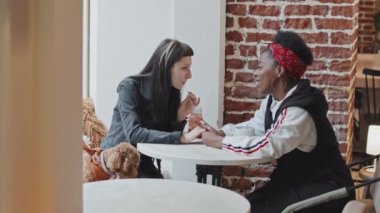 Orta boy beyaz kadın ve Afro-Amerikan kız arkadaşı kafeteryada oturmuş sohbet ederken sevimli köpekleri sabırla onların yanında bekliyor.