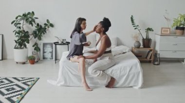 Beyaz güzel bir kadının yatakta oturup Afrika kökenli Amerikalı kız arkadaşına yüz kremi sürdüğü geniş açılı bir çekim.