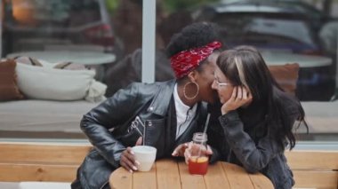 Kafe 'nin dışındaki masada oturan ve kafkas kız arkadaşıyla flört eden mutlu bir Afro-Amerikalı kadının orta boy fotoğrafı.