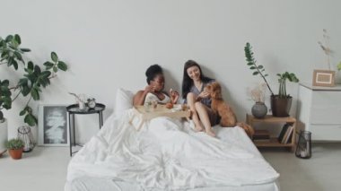 Afrika kökenli Amerikalı ve Kafkas kadınlarının sabahları yatakta dinlenip, kahvaltı yaparken sevimli bir köpek yavrusu besledikleri yavaş çekim.