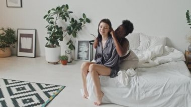 Afro-Amerikalı kız arkadaşı saçlarını sevgiyle tararken beyaz kadının yatakta oturup kahkahalarla güldüğü el kamerasıyla çekilmiş.