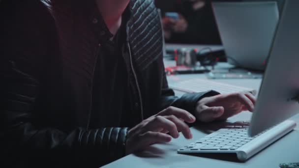 ハンドヘルド追跡ショットアップの若い男性ハッカーでジャケットと帽子に座ってデスクとコーディングでコンピュータ上の暗い隠れ家 — ストック動画