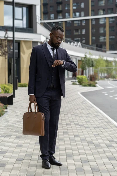Elegáns üzletember kézitáskával nézi a karórát, miközben az úton áll — Stock Fotó
