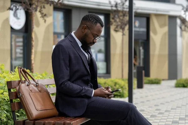 Młody przedsiębiorca afrykańskiego pochodzenia smsy w smartfonie podczas odpoczynku na ławce — Zdjęcie stockowe