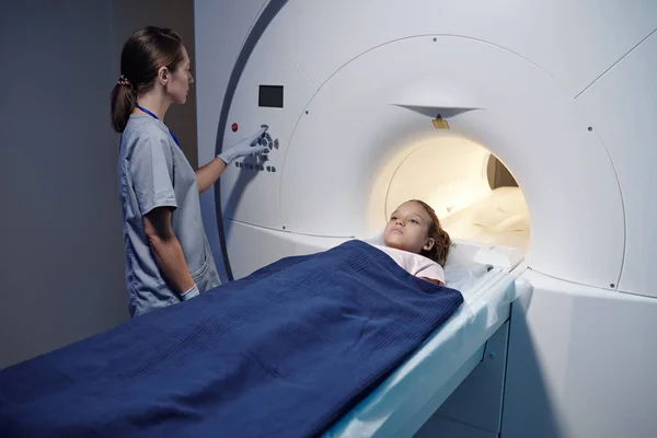 Niña acostada en la mesa de la máquina de escaneo de resonancia magnética mientras el radiografo presiona el botón — Foto de Stock