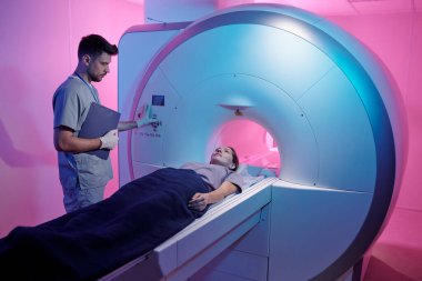 Üniformalı genç doktor hastayla MRI tarama makinesinin başlatma düğmesine basıyor