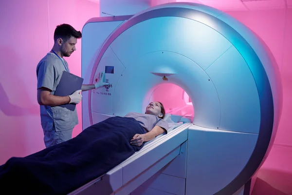 Joven médico en uniforme pulsando el botón de inicio de la máquina de escaneo mri con paciente — Foto de Stock