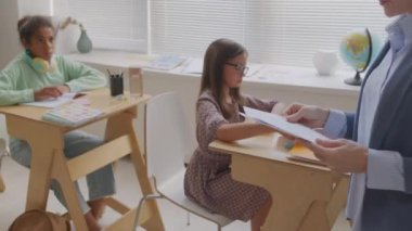 Sınıfta sıraların arasında yürüyen ve ortaokul öğrencilerine test kağıtları veren tanınmayan bayan öğretmenin el bilgisayarı takibi.
