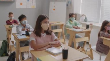 Sınıfta maske takan ve ders sırasında el kaldıran çok ırklı ortaokul öğrencilerinin orta boy fotoğrafları.