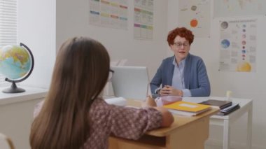 Orta okul çocuklarıyla konuşurken, kısa kıvırcık saçlı, masa başında oturan ve dizüstü bilgisayar kullanan neşeli bayan öğretmenin el kameralı görüntüsü.