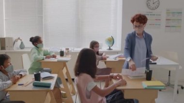 Yüz maskeli kadın öğretmenin, kız ve erkek öğrencilere test kağıtları dağıtırken, aynı zamanda yüz maskesi takıp, sınıftaki masalarda otururken elde çekilmiş bir fotoğrafı.
