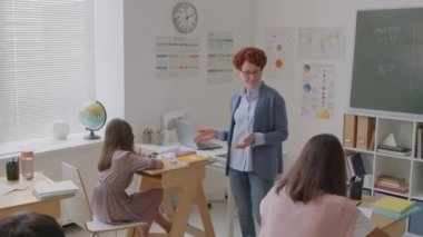 Orta okul sınıfındaki kısa kıvırcık saçlı neşeli bayan öğretmenin el kamerasıyla çekilmiş fotoğrafı ve çocuklardan test kağıtları ya da ödevleri topluyor.