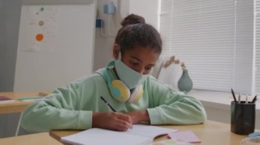 Sınıfta maske takmış, ders sırasında not defterine yazı yazan 11 yaşındaki Afro-Amerikan bir kızın el kamerası görüntüsü.