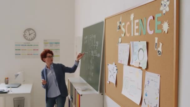 黒板の横に立つ女性科学教師の様子を手に取り 学生に情報を説明する 掲示板に掛けられたポスターや写真 — ストック動画