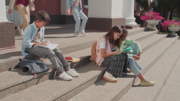 中学校の校舎の階段に座っている子供たちの手持ち撮影 彼らは友達とおしゃべりをしたり宿題をしたりタブレットを使っています — ストック動画