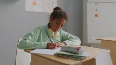 Sınıfta masasına oturmuş ders sırasında not defterine yazı yazan 11 yaşındaki Afro-Amerikan bir kızın el kamerası görüntüsü.