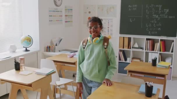 ハンドヘルドトラッキングポートレートショットの幸せなアフリカ系アメリカ人11歳の女の子身に着けているバックパックとヘッドフォン周り彼女の首笑顔のためにカメラのために現代的な中学校の教室 — ストック動画