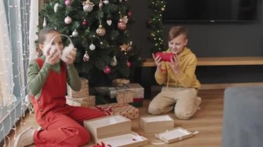 Mutlu sürpriz kardeşler evlerinde güzel bir Noel ağacının altında oturmuş, erkek için kırmızı taşınabilir hoparlör, kız için kürk kulaklık ve eldivenleri olan hediye kutularını açıyorlar.