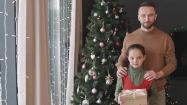 Şefkatli bir babanın PAN portresi ve elinde hediye kutusuyla içeride dekore edilmiş Noel ağacının yanında kameraya gülümseyen 10 yaşındaki güzel kızı.