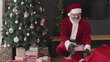 Neşeli Noel Baba, modern ev ya da apartmandaki süslü Noel ağacının altına hediyelerle dolu bir kutu koyuyor.
