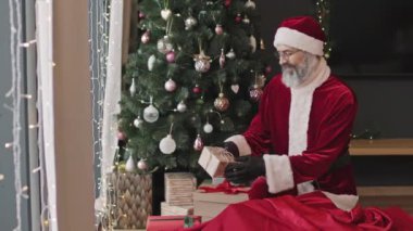 Noel Baba 'nın kırmızı kostümlü neşeli yaşlı adam portresi süslenmiş Noel ağacının yanında duruyor ve bir sürü hediye kutusu var.