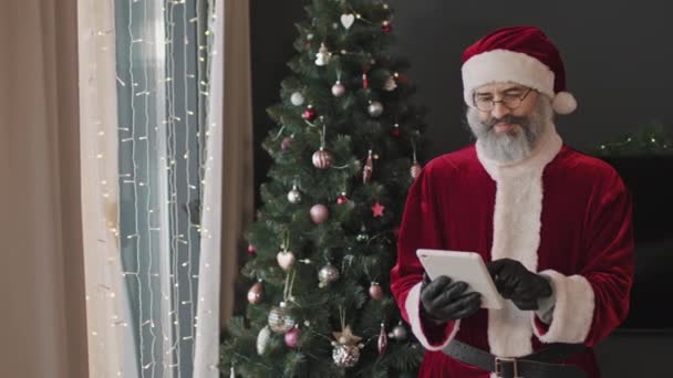 中Pan慢速画面 身穿红色圣诞老人服装的快乐成年人站在美丽的圣诞树旁 手持数码平板电脑 摆出相机的姿势 — 图库视频影像