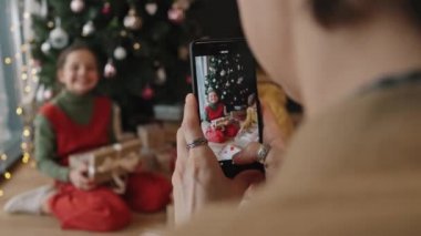 Omuzlarının üstünden, çocuklarının Noel hediyeleriyle fotoğraf çeken bir kadının güzel dekore edilmiş Noel ağacının altında, rahat oturma odasında otururken.