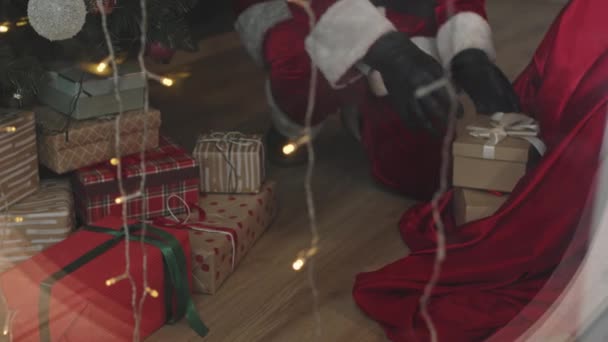 黒い革の手袋で認識できないサンタのパンニングショットクリスマスツリーの下に多くのギフトボックスを置く — ストック動画