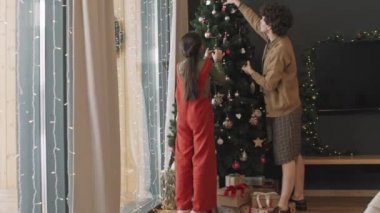 Anne ve kızının rahat oturma odasında Noel ağacını süslediği yavaş bir PAN çekimi.