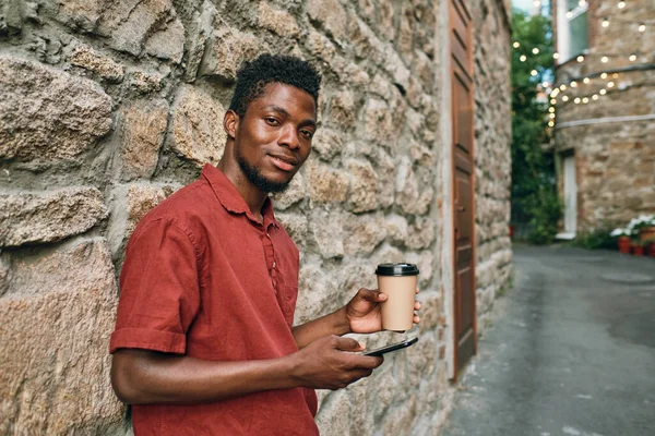 Gelukkig jonge Afrikaanse man in casualwear scrollen in smartphone en het hebben van drinken — Stockfoto