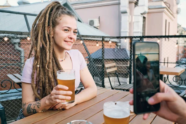 Chica feliz con rastas sosteniendo un vaso de cerveza mientras posan para un amigo con teléfono inteligente —  Fotos de Stock