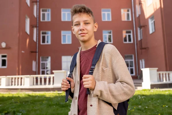 Menino feliz com mochila em pé no gramado verde contra a escola — Fotografia de Stock