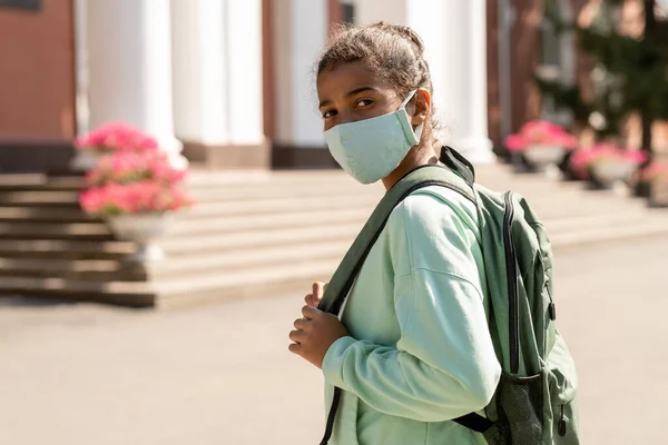 Gadis sekolah ras campuran yang lucu dengan topeng pelindung pergi ke sekolah — Stok Foto