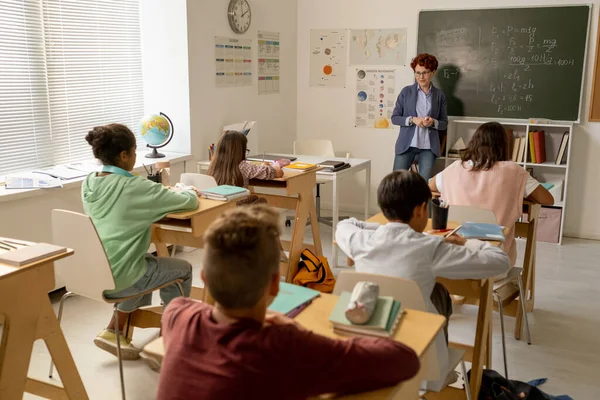 Joven profesor de pie junto a la pizarra frente a los escolares interculturales — Foto de Stock