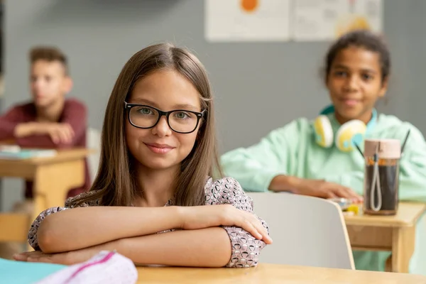 Cute uczennica w okularach patrząc na ciebie przez biurko — Zdjęcie stockowe