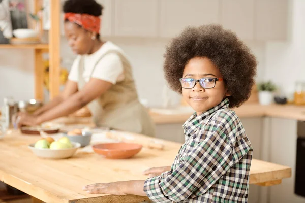 Niedlicher kleiner Junge steht am hölzernen Küchentisch gegen seine Mutter — Stockfoto