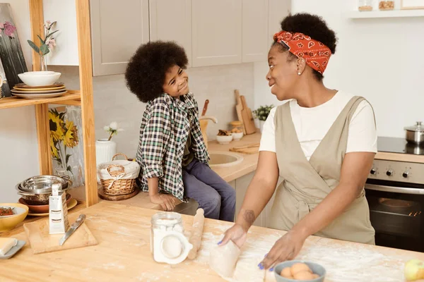 Feliz lindo niño sentado junto a la mesa de la cocina mientras su mamá cocina pastelería —  Fotos de Stock