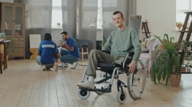 Tekerlekli sandalyede kameraya poz veren gülümseyen beyaz bir adamın yavaş yavaş portresi. Bir grup gönüllü evinde mobilyaları monte etmeye ya da tamir etmeye yardım ediyor.