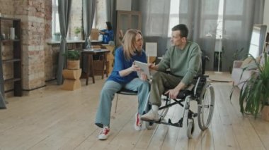 Tekerlekli sandalyedeki engelli genç adamın dijital tablete internet kurmasına yardım eden ve sonra kameraya gülümseyen bir kadın portresi.