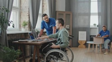 Özürlü genç bir adama evinde yardım eden, dizüstü bilgisayara internet kuran ve karton kutulardan giysileri çıkaran gönüllülerin yavaş çekimleri.