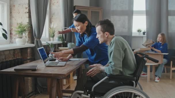 跟踪多种族志愿者小组的慢镜头 该小组向坐在轮椅上的残疾年轻人解释如何在他的电脑上使用互联网 同时在他的公寓里安装无线路由器 — 图库视频影像