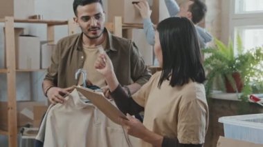 Asyalı genç kadın liderin yavaş çekimleri ellerinde panoyla stüdyoda bağışlanan kıyafetleri düzenlerken ve iş arkadaşlarına emir verirken.