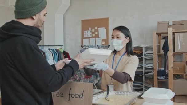 一名戴着面具的亚洲女性志愿者站在室内餐桌旁 用塑料容器向有需要的人提供食物 — 图库视频影像