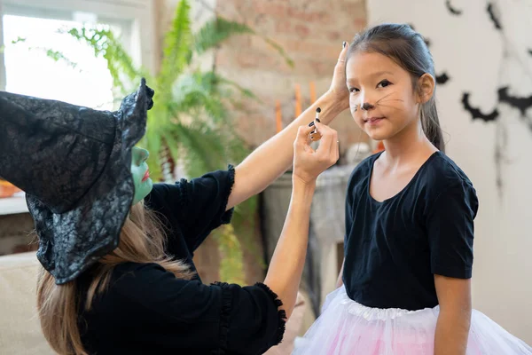 Jovem fêmea em chapéu de bruxa e pulôver preto aplicando maquiagem halloween no rosto da menina bonito — Fotografia de Stock