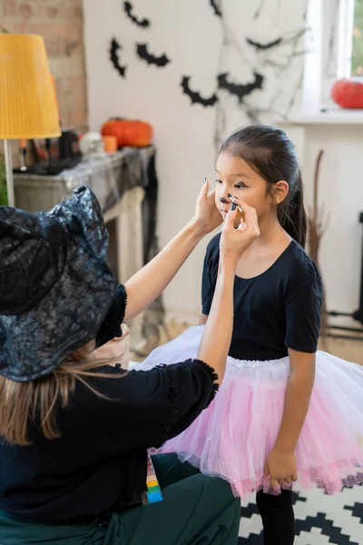 Jonge vrouw in zwart halloween kleding het aanbrengen van make-up op gezicht van klein meisje — Stockfoto