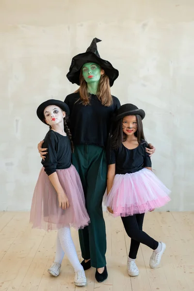 Jovem bruxa com o rosto pintado de verde abraçando duas meninas — Fotografia de Stock