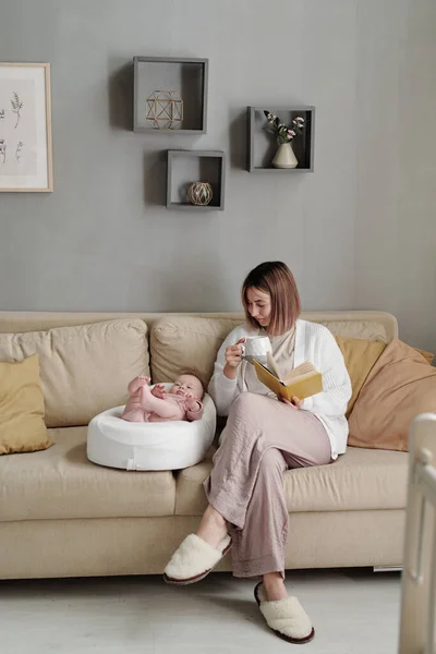 Mujer joven con taza de té y libro sentado en el sofá por su hija bebé — Foto de Stock