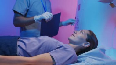 Heyecanlı bir kadının tarama masasında MRI incelemesi için tüpün içine girerken yavaş yavaş yan görüntüsü. Doktor modern ekipmanların kontrol panelindeki düğmelere basıyor