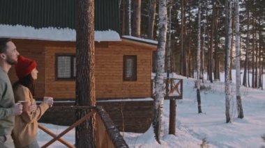 Elinde sıcak çay bardakları olan mutlu erkek ve kadının el kamerasıyla ve kulübelerinin verandasından kış manzarasının keyfini çıkarırken.