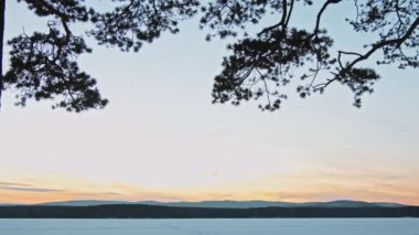 Kışın gün batımında kar, orman ve tepelerle kaplı donmuş gölün durgun görüntüsü.